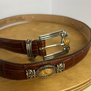 Puede incluir: Un cintur&oacute;n de cuero marr&oacute;n con hebilla plateada y detalles decorativos. El cintur&oacute;n tiene un patr&oacute;n texturizado similar al de un cocodrilo. La hebilla y los elementos decorativos tienen un centro dorado.