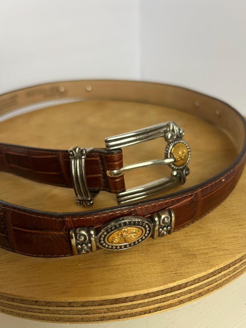 Puede incluir: Un cintur&oacute;n de cuero marr&oacute;n con hebilla plateada y detalles decorativos. El cintur&oacute;n tiene un patr&oacute;n texturizado similar al de un cocodrilo. La hebilla y los elementos decorativos tienen un centro dorado.