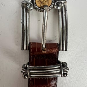 Puede incluir: Un cintur&oacute;n de cuero marr&oacute;n con una hebilla plateada. La hebilla presenta un medall&oacute;n dorado con una imagen de perfil y detalles plateados ornamentados. El cintur&oacute;n tiene un patr&oacute;n texturizado similar al de un cocodrilo.