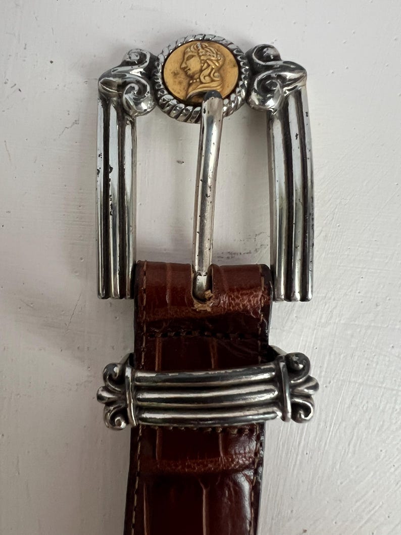 Puede incluir: Un cintur&oacute;n de cuero marr&oacute;n con una hebilla plateada. La hebilla presenta un medall&oacute;n dorado con una imagen de perfil y detalles plateados ornamentados. El cintur&oacute;n tiene un patr&oacute;n texturizado similar al de un cocodrilo.