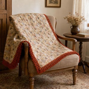 May include: A rectangular throw blanket featuring a dense, multi-colored floral pattern in shades of cream, tan, and brown. The blanket is finished with a thick, textured rust-colored fringe border along the edges, creating a decorative trim.