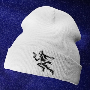 May include: White beanie with a black embroidered design of a person running with a gun. The design is on the front of the beanie.