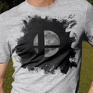 Può includere: Una maglietta grigia con una grafica in bianco e nero del logo di Super Smash Bros. con una luna al centro.