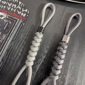 May include: Two gray paracord lanyards with braided sections and looped ends. One lanyard has black metal rings, the other has silver metal rings. A playing card box with "HA HA HA" text is visible in the background.