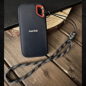 May include: A navy blue Sandisk external hard drive with a perforated texture and a red accent. It has a black and white lanyard attached. The hard drive is on a wooden surface.