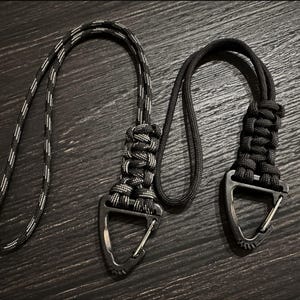 May include: Two black and gray paracord lanyards with metal carabiner clips. One lanyard has a black and white striped pattern, while the other is solid black. The lanyards are designed for attaching keys or other small items.