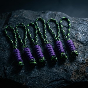 May include: Six zipper pulls made from black and neon green paracord, each featuring a thick, braided purple cord section at the base and a small knotted end. The pulls are arranged in a row, each with a looped top for attachment to a zipper.