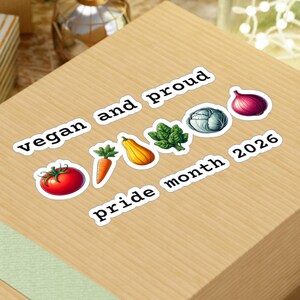 Könnte beinhalten: Eine Holzgeschenkbox mit einem hellgrünen Band, verziert mit Aufklebern mit der Aufschrift "vegan and proud" und "pride month 2026". Die Aufkleber zeigen Illustrationen von Gemüse. Die Box ist von festlichen Dekorationen umgeben.