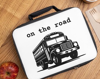 Puede incluir: Una bolsa de almuerzo blanca con una cremallera negra y un asa negra. La bolsa tiene una ilustración en blanco y negro de un autobús escolar con el texto "on the road" impreso en la parte delantera.