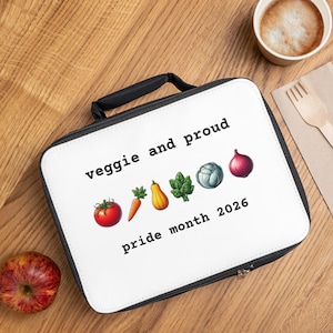 May include: A white lunch bag with a black handle and zipper, featuring the text "veggie and proud" and "pride month 2026." The bag is decorated with illustrations of vegetables. An apple, a cup of coffee, and a fork are on a wooden surface.