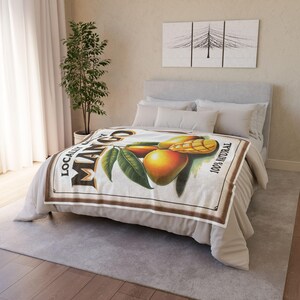 Puede incluir: Una manta blanca con una ilustración de estilo vintage de mangos y el texto "LOCAL MANGO 100% NATURAL". La manta está sobre una cama con ropa de cama y almohadas blancas.