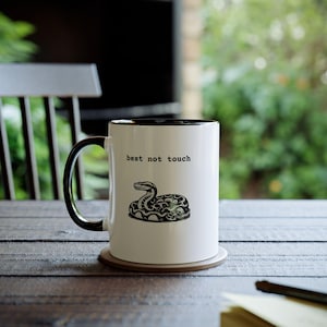May include: White ceramic coffee mug with a black rim and handle. The mug has a black and white illustration of a snake with the text "best not touch" printed on the front.