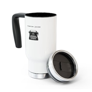 May include: White travel mug with a black handle and lid. The mug has a black and white illustration of a typewriter and the text "creative juices".