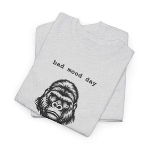 May include: Two light gray sweatshirts are stacked. The top shirt features the text "bad mood day" above a black and white illustration of a gorilla's face. The sweatshirts are folded.