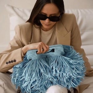 May include: A light blue clutch bag featuring a structured frame and a dense exterior covered in long, thin, matching blue fringe strips. The bag has a woven crochet-style top handle and a rectangular shape with a voluminous, textured appearance.