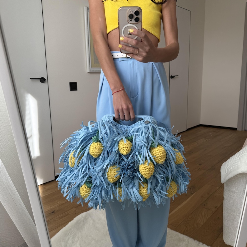 May include: A light blue crochet handbag featuring long, dangling fringe strips and multiple small, yellow, textured pineapple-shaped embellishments attached throughout the exterior. The bag has a rounded shape with a sturdy, integrated crochet handle.