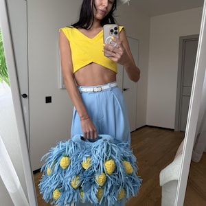May include: A light blue crochet handbag featuring long fringe strips and multiple small, yellow, textured pineapple-shaped embellishments attached throughout the exterior. The bag has a structured handle and a soft, yarn-based construction.