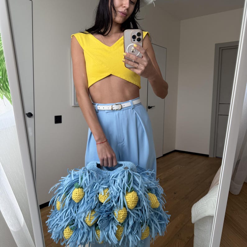 May include: A light blue crochet handbag featuring long fringe strips and multiple small, yellow, textured pineapple-shaped embellishments attached throughout the exterior. The bag has a structured handle and a soft, yarn-based construction.