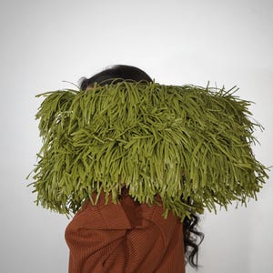 May include: A rectangular clutch bag covered in long, thin, olive green fringe strips. The bag is held horizontally, obscuring the upper portion of the figure. The fringe is dense and uniform, creating a textured, shaggy appearance across the surface.