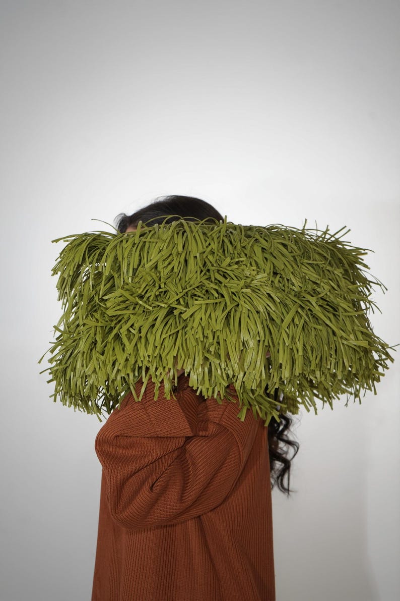 May include: A rectangular clutch bag covered in long, thin, olive green fringe strips. The bag is held horizontally, obscuring the upper portion of the figure. The fringe is dense and uniform, creating a textured, shaggy appearance across the surface.