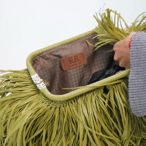 May include: A lime green clutch bag featuring long, thin fringe strips covering the exterior. The bag has a metal frame closure and an interior lining with a brown leather-like patch embossed with the letters RB in the center of the opening.