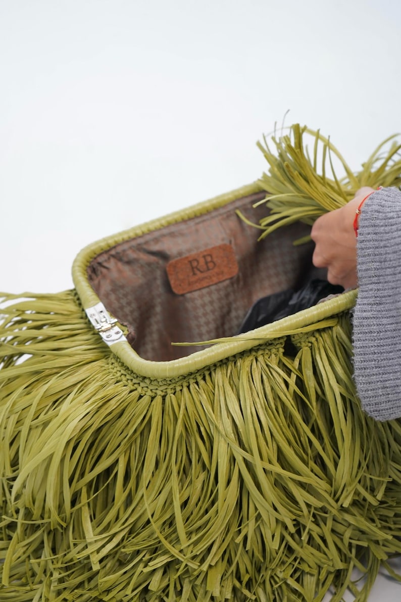 May include: A lime green clutch bag featuring long, thin fringe strips covering the exterior. The bag has a metal frame closure and an interior lining with a brown leather-like patch embossed with the letters RB in the center of the opening.