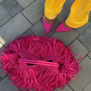 Könnte beinhalten: Eine leuchtend pinkfarbene Clutch-Tasche mit Fransen. Die Tasche besteht aus einem gewebten Material und hat einen langen, dünnen Griff. Die Tasche liegt auf einer grauen Ziegeloberfläche. Das Bild zeigt auch gelbe Hosen und pinkfarbene Schuhe mit spitzer Zehenpartie.