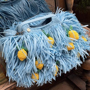 May include: A light blue crocheted handbag featuring long fringe detailing and multiple small, yellow, textured lemon-shaped embellishments with green leaves. The bag includes a sturdy handle and a small circular tag with the letters RB.