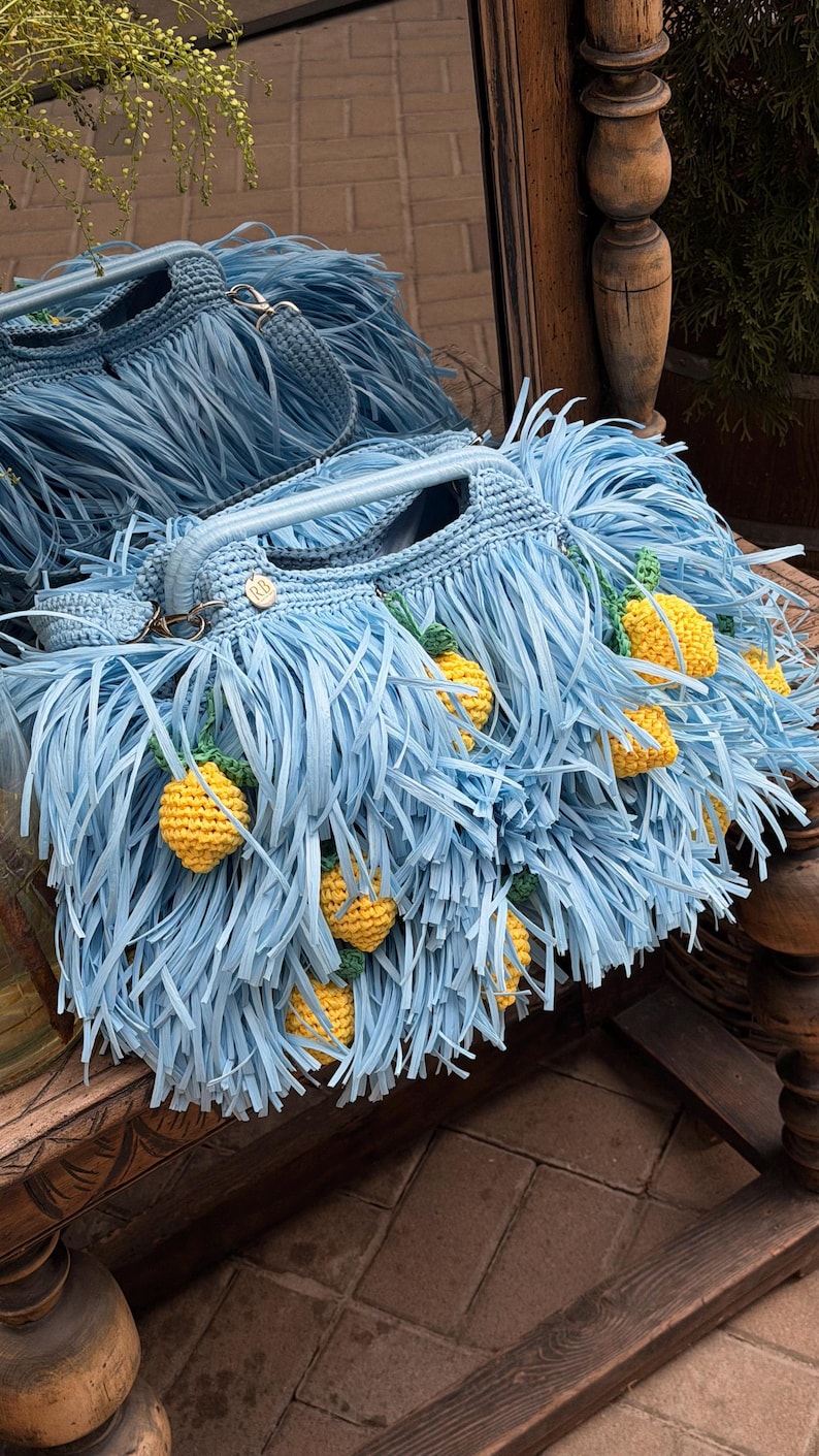 May include: A light blue crocheted handbag featuring long fringe detailing and multiple small, yellow, textured lemon-shaped embellishments with green leaves. The bag includes a sturdy handle and a small circular tag with the letters RB.