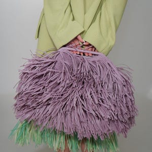 May include: A lavender fringed handbag with a woven top is held in front of a person wearing a light green sweater. The handbag has a large amount of fringe. Below the handbag is a skirt with green and white fringe.