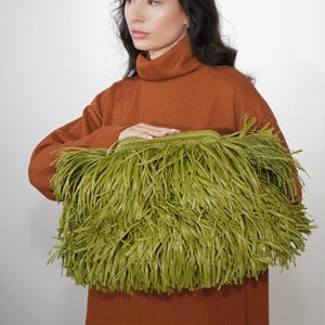 May include: A large, rectangular clutch bag in a vibrant olive green color. The exterior is covered in long, thin, dangling strips of material that create a dense, shaggy fringe texture. The top of the bag features a solid, crocheted green border.