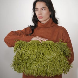 May include: A rectangular clutch bag in a vibrant olive green color, featuring a dense exterior layer of long, thin, fringed strips. The bag has a structured, woven top frame and a secure clasp closure, providing a textured and layered appearance.