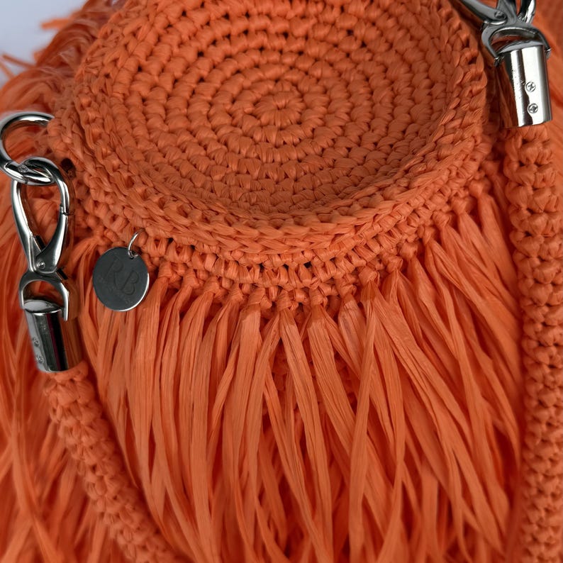 May include: A bright orange crocheted bag featuring a circular base with dense, long fringe detailing. The bag includes a braided strap attached with silver-toned metal lobster clasps and a small, round metal charm engraved with the letters RB.