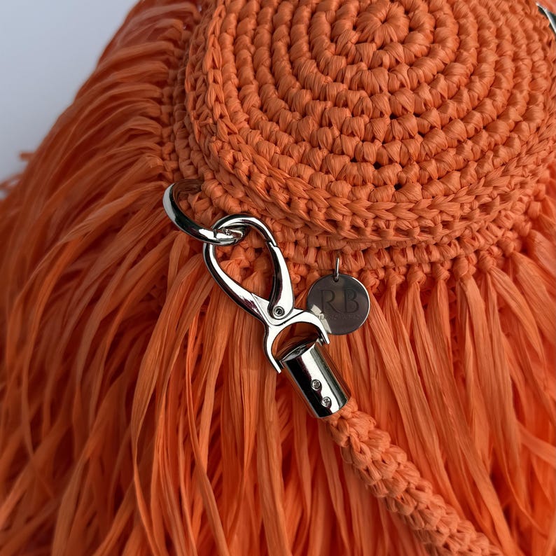 May include: A bright orange crocheted bag featuring a dense fringe texture. The bag includes a silver-toned metal lobster clasp attached to a circular ring and a matching braided strap. A small, round silver-toned metal tag with the letters RB is attached.