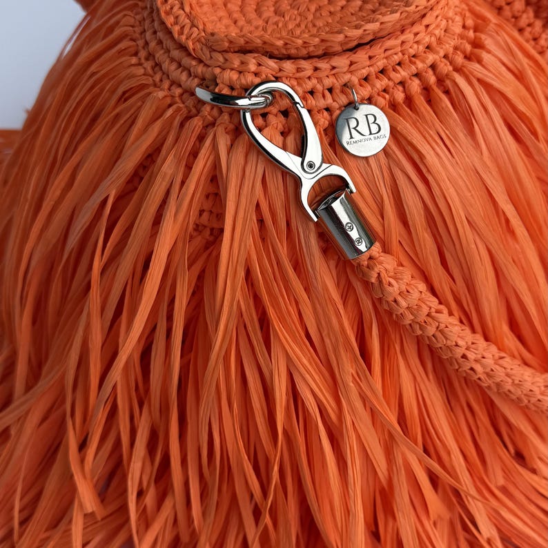 May include: A bright orange crocheted bag featuring long, thin fringe strands. A silver-toned metal lobster clasp attaches a braided strap to the bag. A small, round silver metal tag is attached, engraved with the letters RB and REMNOVA BAGS.
