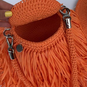 May include: A bright orange crocheted handbag featuring a circular flap, a braided shoulder strap attached with silver-toned metal lobster clasps, and long fringe detailing. A small, round silver-toned metal tag with the letters RB is attached.