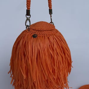May include: A bright orange handbag featuring a rounded, crocheted top section and a dense, fringed body made of long, thin strips. The bag includes a braided handle attached with silver-toned metal clasps and a small, circular silver-toned metal charm.