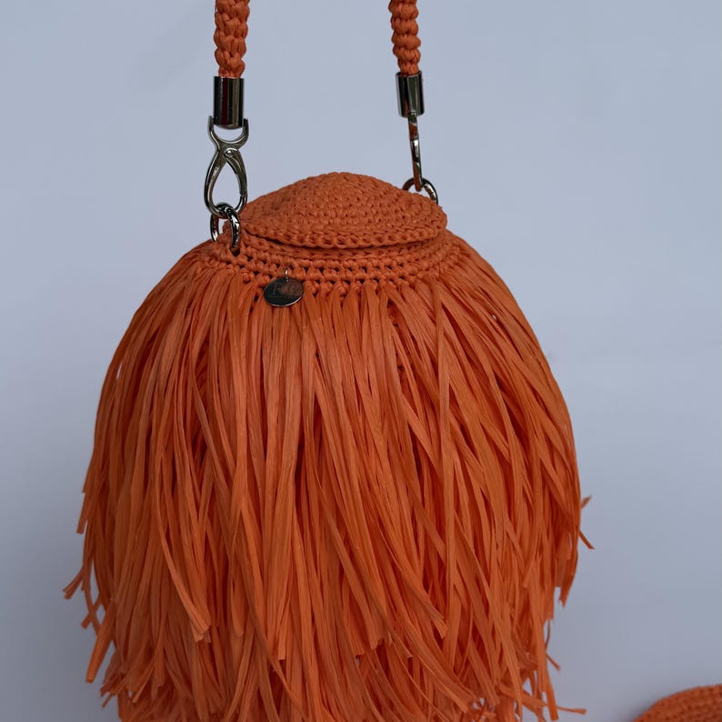 May include: A bright orange handbag featuring a rounded, crocheted top section and a dense, fringed body made of long, thin strips. The bag includes a braided handle attached with silver-toned metal clasps and a small, circular silver-toned metal charm.