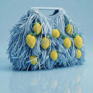 May include: A light blue handbag featuring a dense fringe texture and multiple small, crocheted yellow lemon motifs with green leaves attached to the exterior. The bag includes a rigid, matching light blue top handle and a small circular silver logo tag.
