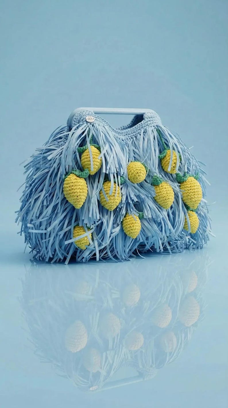 May include: A light blue handbag featuring a dense fringe texture and multiple small, crocheted yellow lemon motifs with green leaves attached to the exterior. The bag includes a rigid, matching light blue top handle and a small circular silver logo tag.