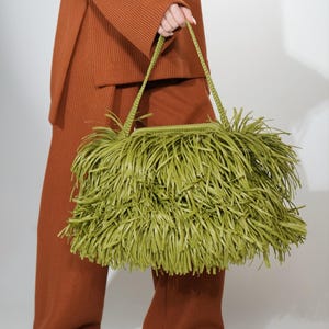 May include: A lime green handbag featuring a dense, layered fringe exterior and a braided shoulder strap. The bag has a rectangular structure and is covered in long, thin strips of material that create a textured, shaggy appearance.