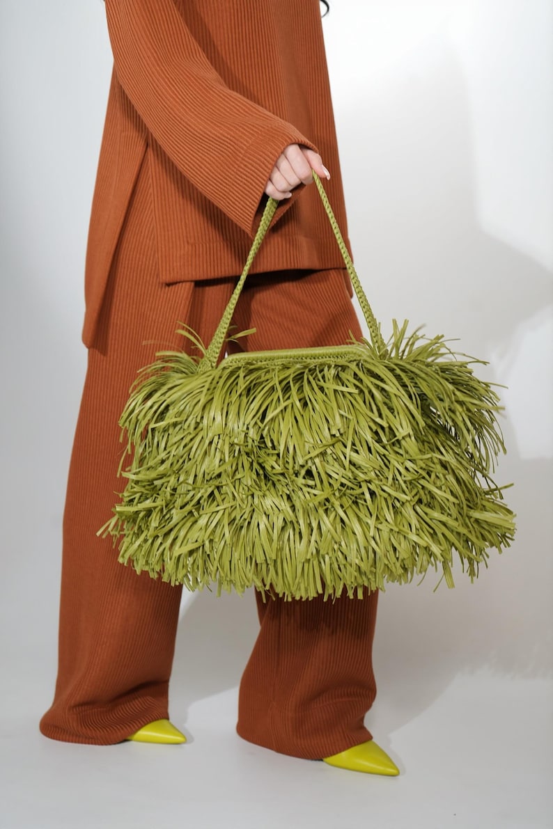 May include: A lime green handbag featuring a dense, layered fringe exterior and a braided shoulder strap. The bag has a rectangular structure and is covered in long, thin strips of material that create a textured, shaggy appearance.