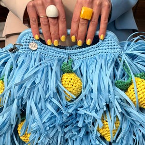 May include: A light blue crocheted handbag featuring long, thin fringe strips and attached yellow crocheted lemon motifs with green leaves. The bag includes a circular silver-toned metal tag engraved with the letters RB.