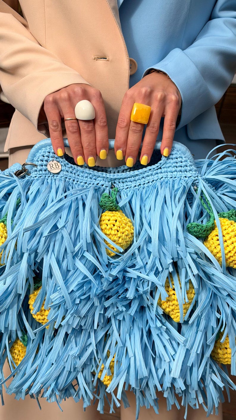 May include: A light blue crocheted handbag featuring long, thin fringe strips and attached yellow crocheted lemon motifs with green leaves. The bag includes a circular silver-toned metal tag engraved with the letters RB.