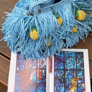May include: A light blue crocheted handbag featuring long fringe and small, yellow, textured lemon-shaped charms hanging from the exterior. The bag has a structured handle and is positioned above an open book displaying stained glass art.