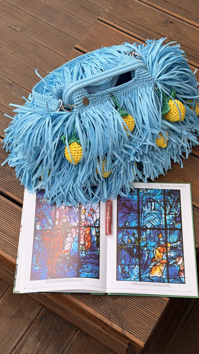 May include: A light blue crocheted handbag featuring long fringe and small, yellow, textured lemon-shaped charms hanging from the exterior. The bag has a structured handle and is positioned above an open book displaying stained glass art.