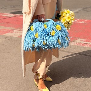 May include: A light blue crochet handbag featuring long fringe and small, yellow, textured lemon-shaped embellishments. The bag has a structured handle and is carried alongside a bouquet of yellow daffodils, wearing beige trousers and yellow heels.