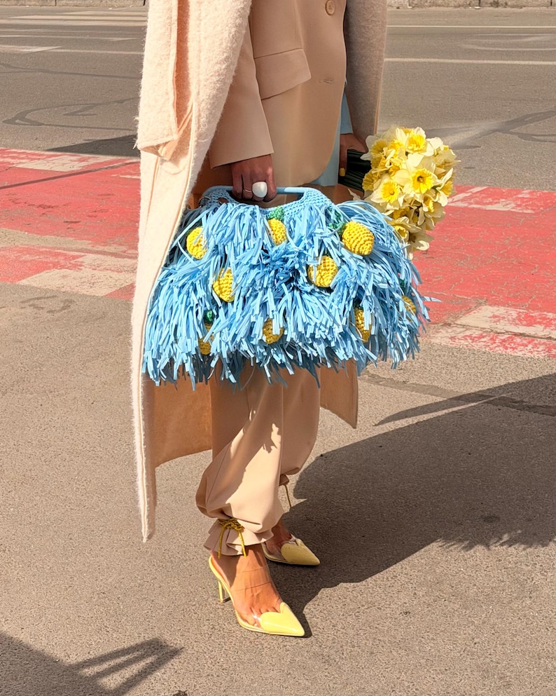 May include: A light blue crochet handbag featuring long fringe and small, yellow, textured lemon-shaped embellishments. The bag has a structured handle and is carried alongside a bouquet of yellow daffodils, wearing beige trousers and yellow heels.