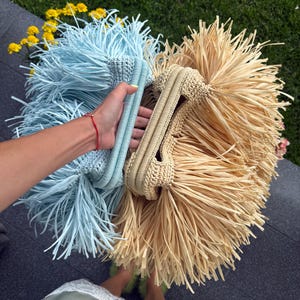 Op de afbeelding: Twee gehaakte handtassen met een stijve frame-sluiting, één in lichtblauw en één in beige. Beide tassen zijn voorzien van lange, dunne, franje-achtige stroken materiaal die naar buiten uitsteken, wat zorgt voor een textuurrijk en volumineus uiterlijk.