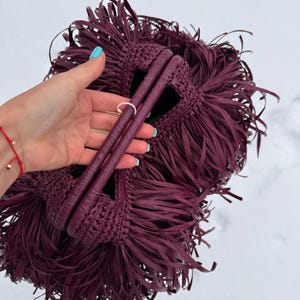 May include: A deep burgundy handbag featuring a dense, layered fringe design covering the entire exterior. The bag is constructed with a crocheted texture and includes two rounded, matching burgundy handles at the top for carrying.